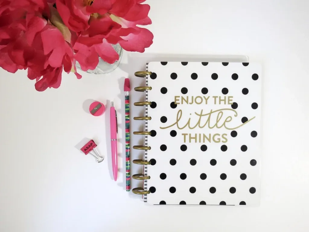A polka-dot planner with a pink pen and flowers on a white desk